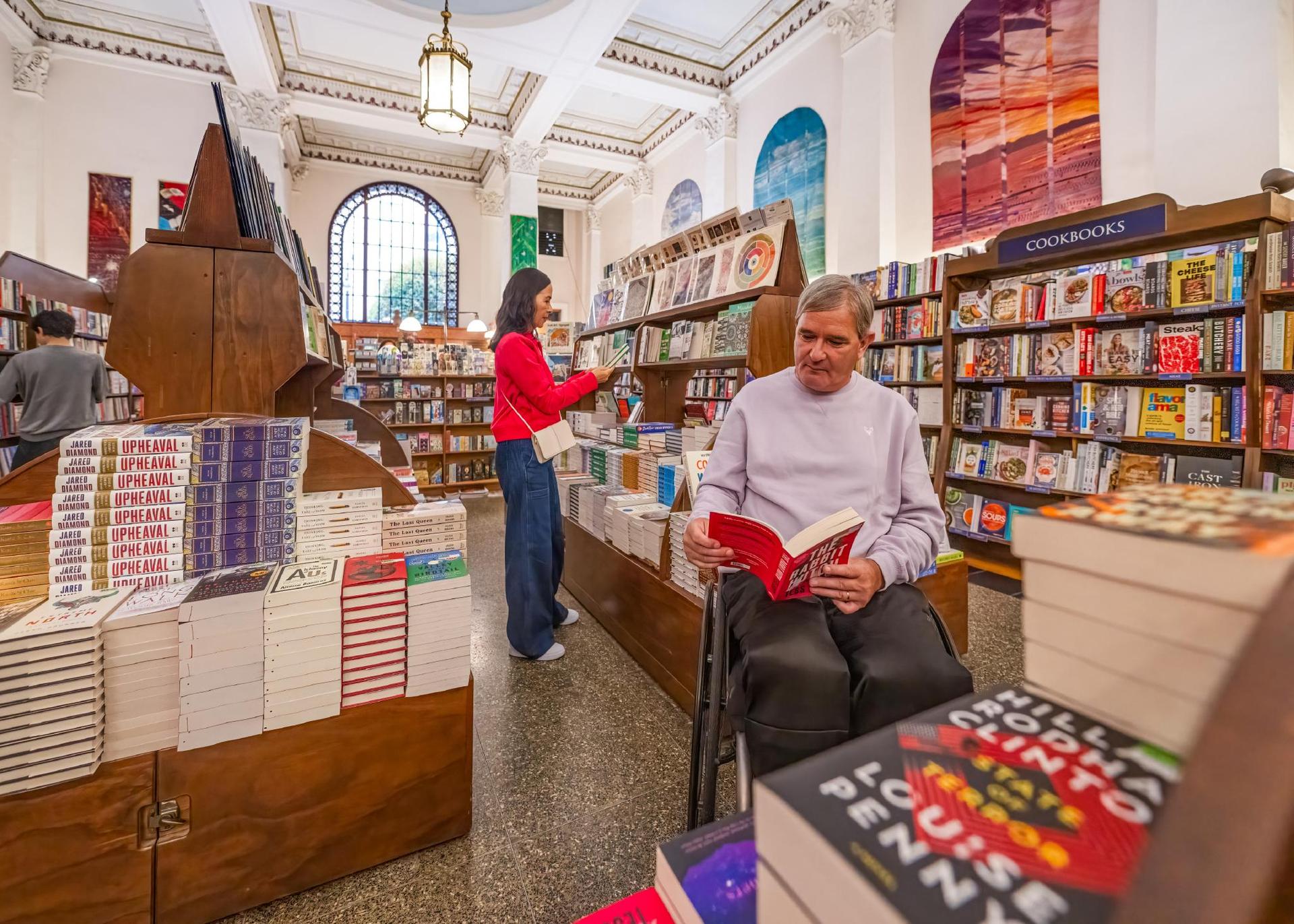 A man in a wheelchair and a woman in a red coat shop for books in Munro's Books, a bookstore in a historic building in Victoria, BC