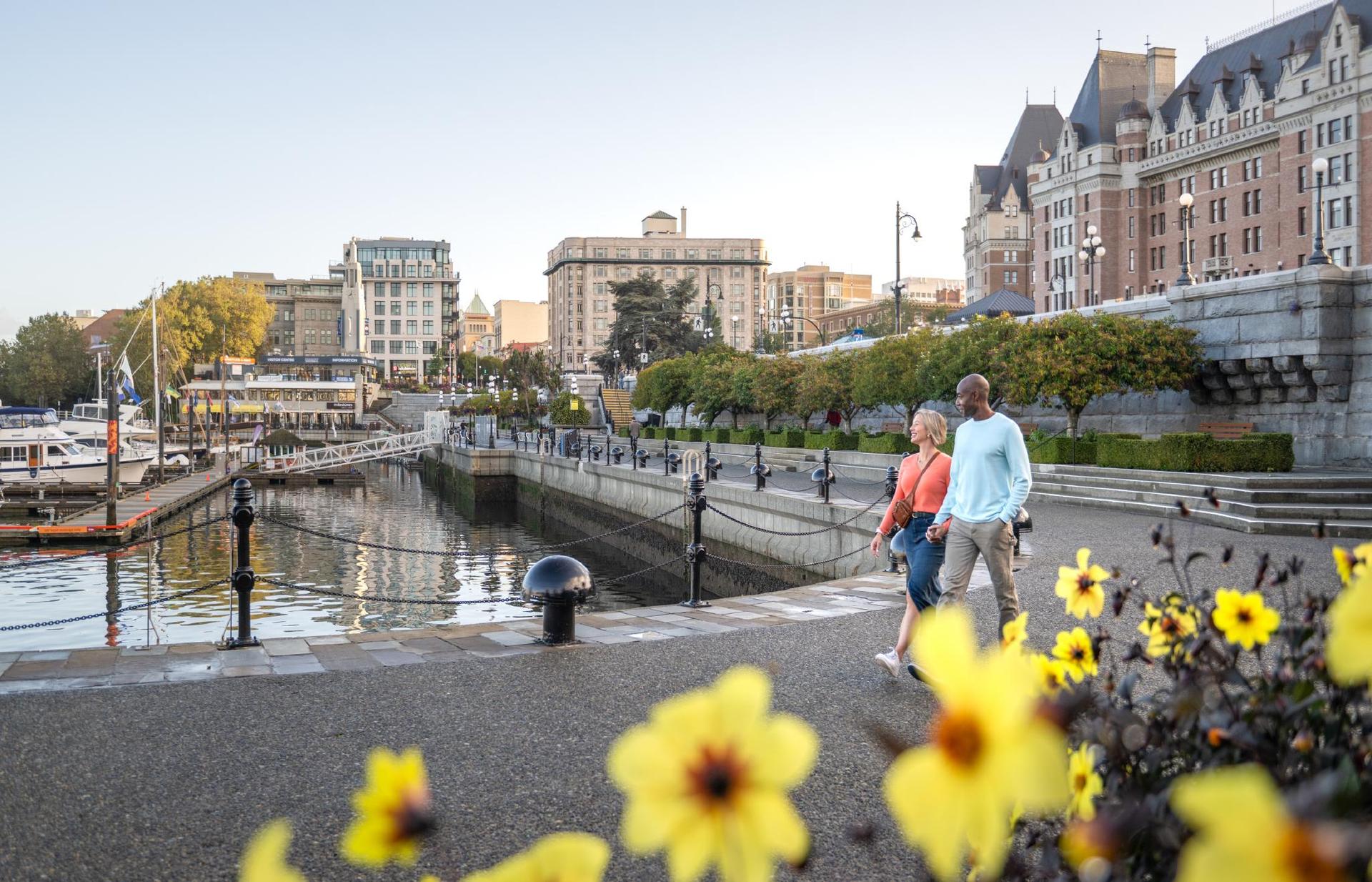 A couple walks along the lower causeway of the Inner Harbour in Victoria, BC as flowers bloom along the harbour.