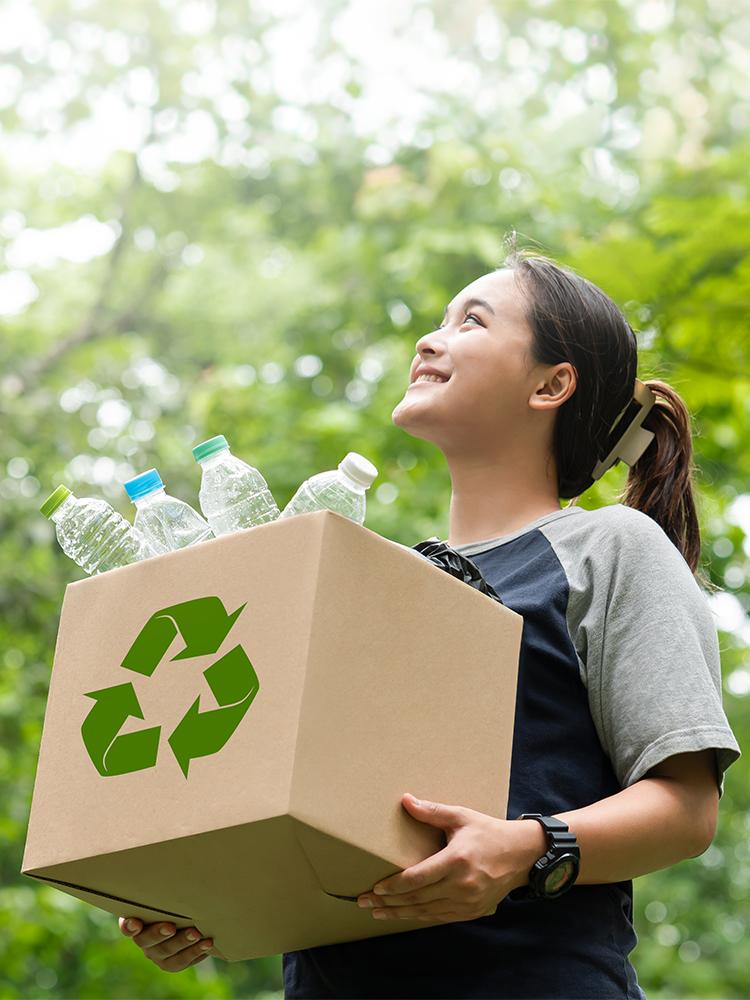 A woman carries a box of recycling through some trees