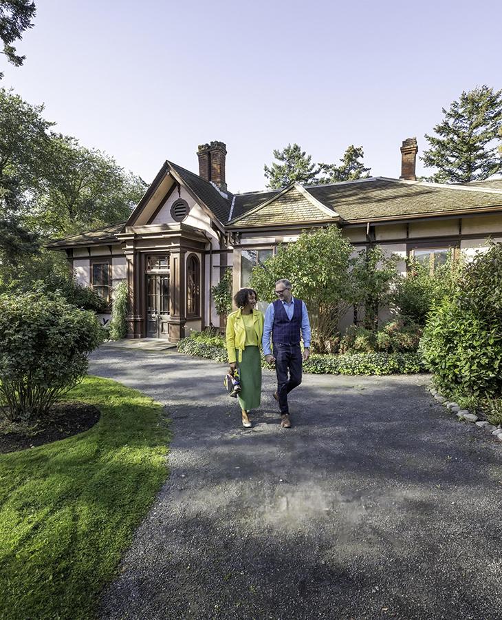 A woman in a green skirt and yellow top walks with a man dressed in blue through the garden at Point Ellice House in Victoria, BC