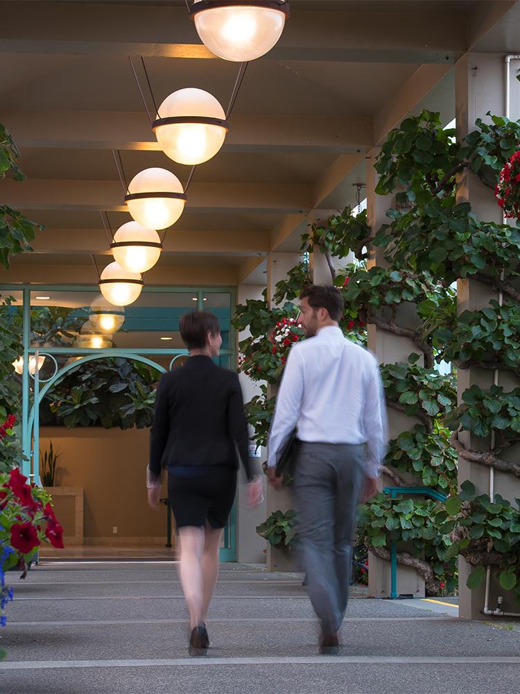 Two professionals, a man in a white shirt and a woman in a skirt and jackets, walk while having a conversation outside at the Victoria Conference Centre in Victoria, BC