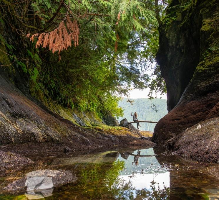 A woman sits on a log over a creek in the rainforest near Victoria, BC