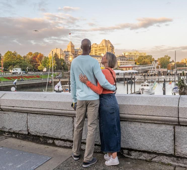 A couple stands with arms around each other looking over Victoria, BC's Inner Harbour during sunrise