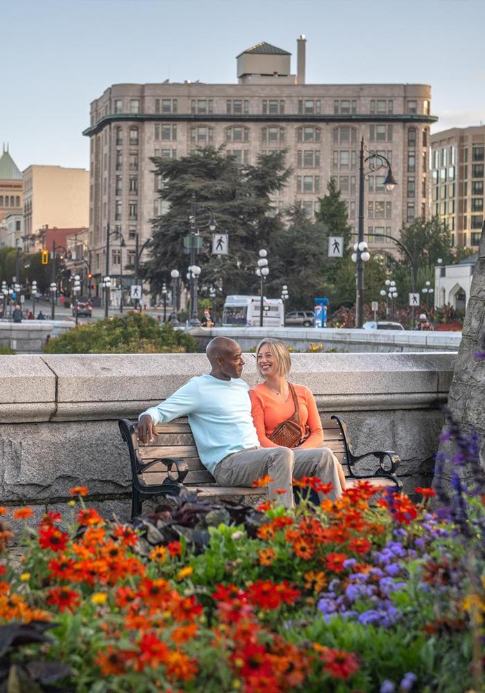 A man in a blue shirt and a woman in an orange shirt sit along the Upper Causeway in front of a garden bed along Victoria, BC's Inner Harbour