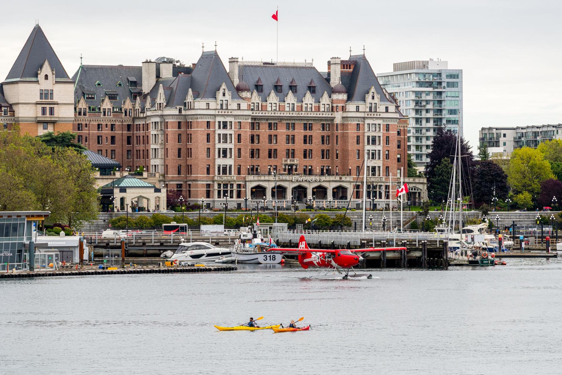 Two kayakers in yellow and orange kayaks paddle Victoria BC's Inner Harbour, backdropped by the Fairmont Empress, while a red Harbour Air Seaplane takes off.