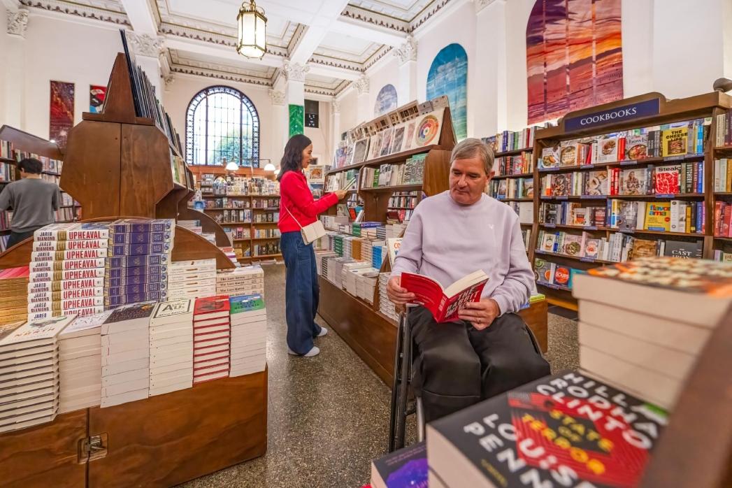 A man in a wheelchair and a woman in a red coat shop for books in Munro's Books, a bookstore in a historic building in Victoria, BC