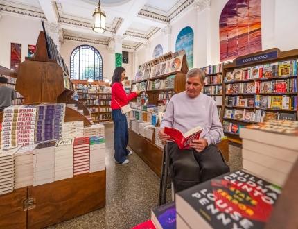 A man in a wheelchair and a woman in a red coat shop for books in Munro's Books, a bookstore in a historic building in Victoria, BC