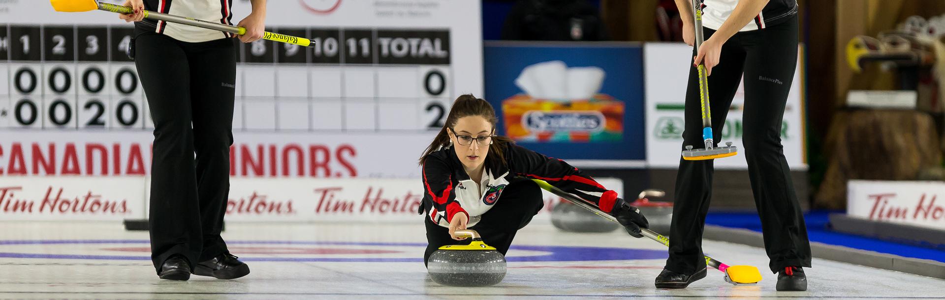 A female curler dressed in black, white, and red curls a yellow rock during a curling competition in Victoria, BC