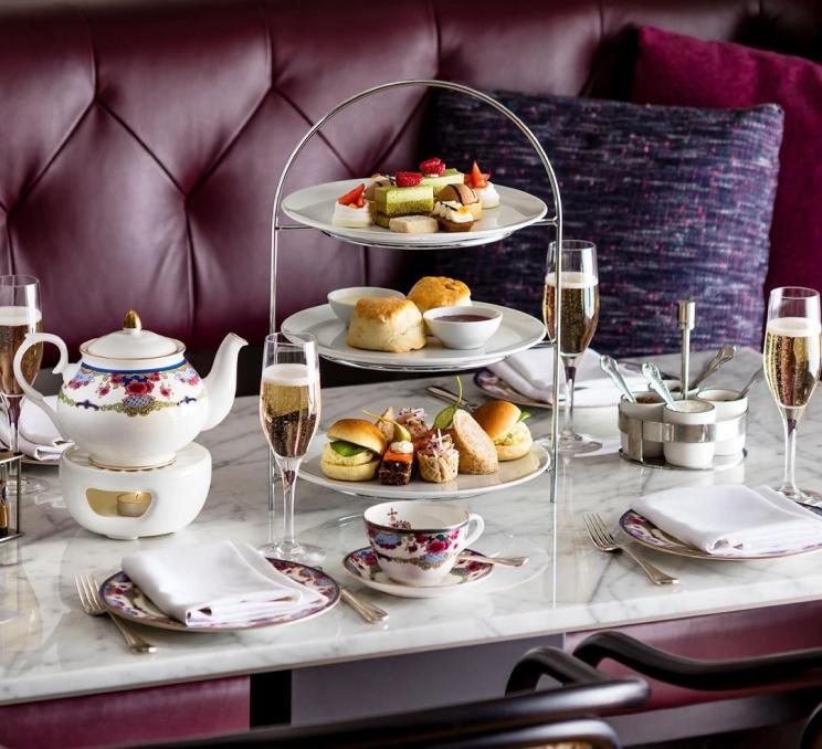An afternoon tea session id displayed with teapots and a three-tier tray of baked goods and sandwiches on a table in front of a purple couch at the Fairmont Empress in Victoria, BC