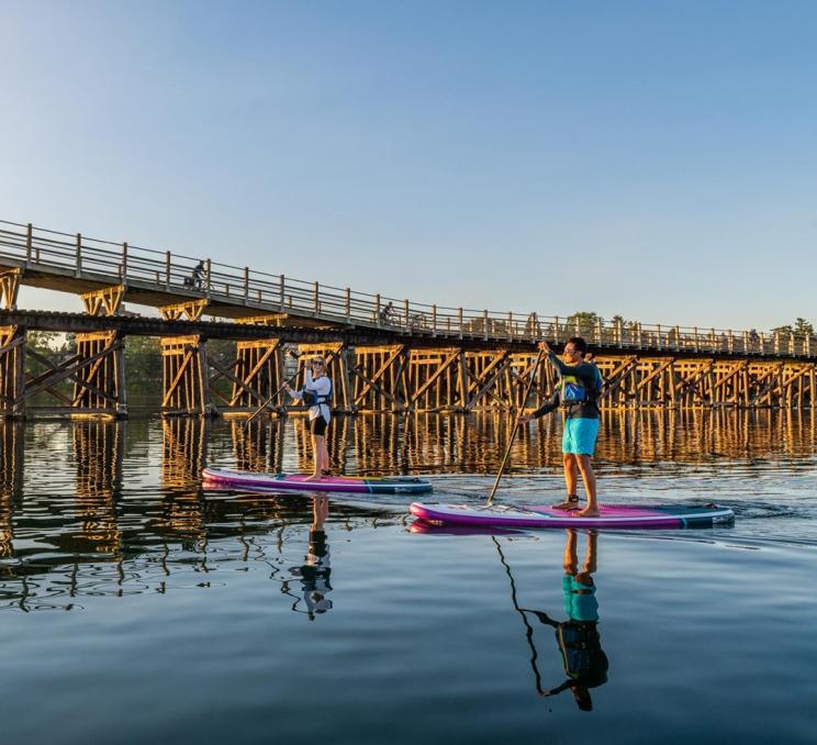 A couple paddle purple paddleboards along the Upper Harbour in front of the wood Selkirk Trestle in Victoria, BC